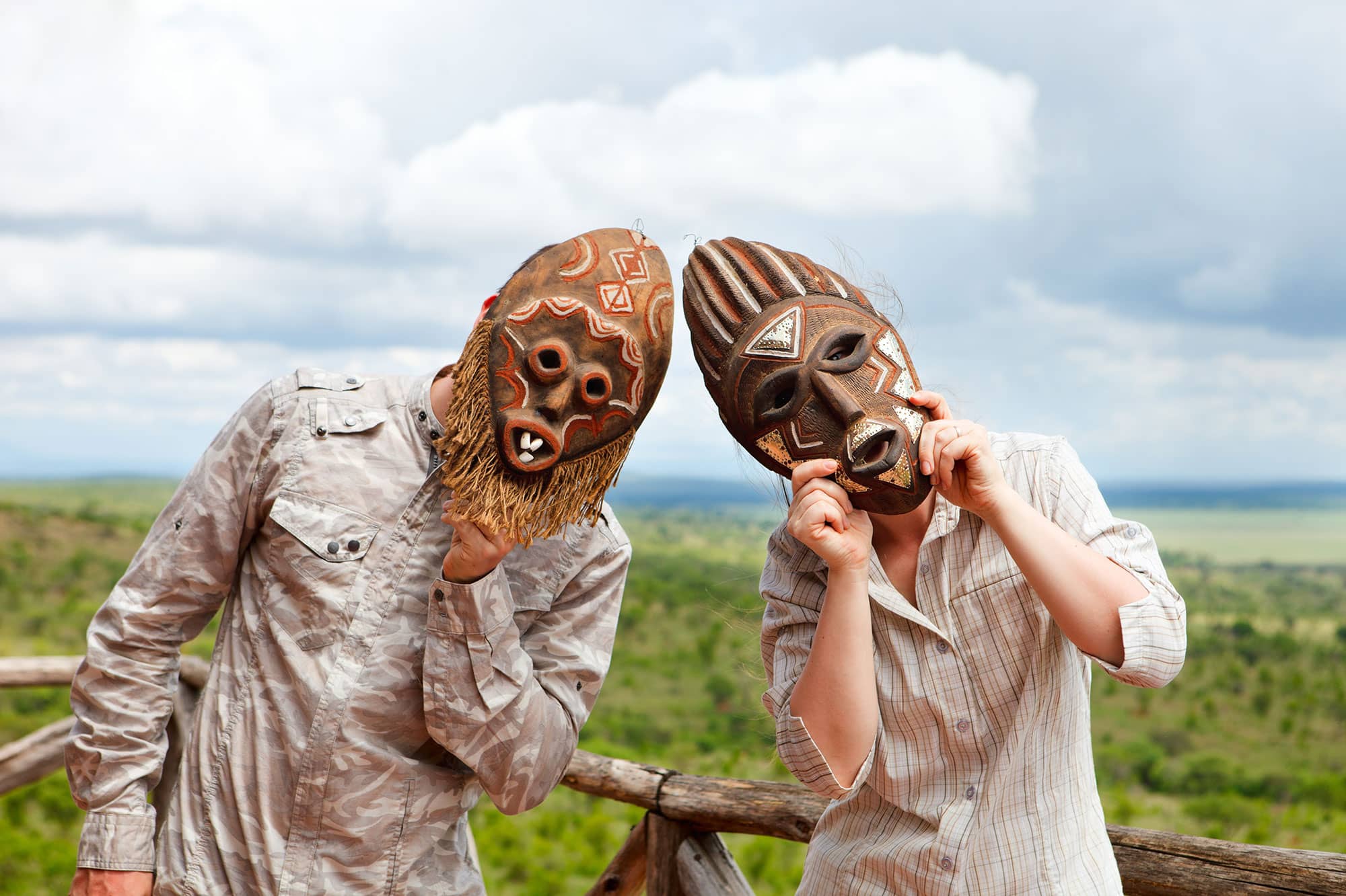 Two tourists hold carved African masks over their faces in front of the Ngorongoro Crater